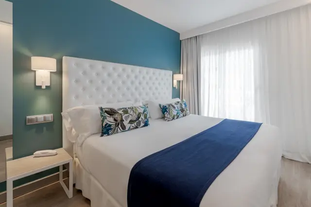 Habitación con cama grande, cabecero blanco acolchado, pared azul turquesa, mesita de noche blanca y cortinas claras