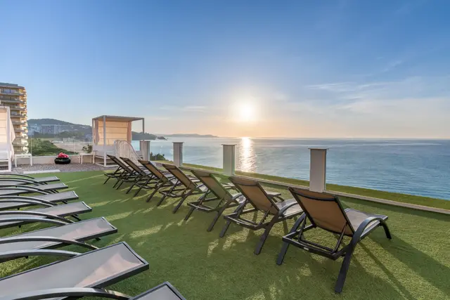 Terraza exterior con césped artificial, varias tumbonas alineadas, barandilla con vistas al mar al atardecer