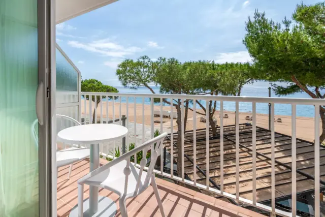 Vista desde el balcón de una suite con mesa y sillas blancas, barandilla blanca, árboles, playa de arena y mar