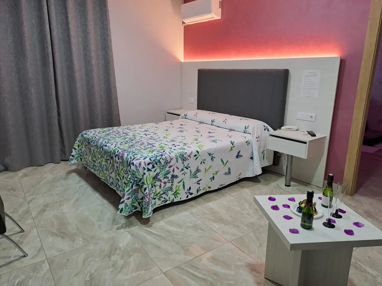 Habitación de hotel con cama doble cubierta por un edredón blanco con estampado floral y mariposas, cabecero gris
