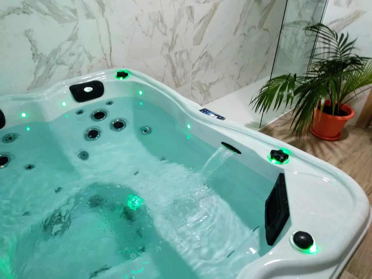 Jacuzzi blanco con agua iluminada en verde, chorros activos y cascada, ubicado en una habitación con paredes de mármol