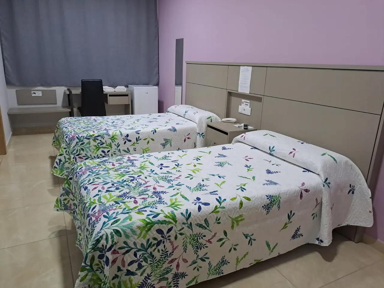 Habitación de hotel con dos camas individuales cubiertas por colchas blancas con estampado floral en tonos verdes
