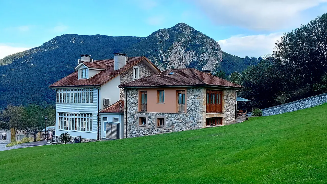 Vista exterior de un edificio de hotel con fachada de piedra y tejado rojo, situado en una ladera verde con montañas
