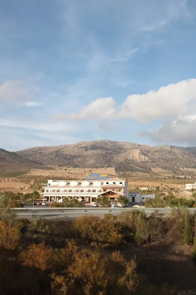 Vista exterior de un hotel de varios pisos con montañas áridas al fondo, bajo un cielo azul con nubes.