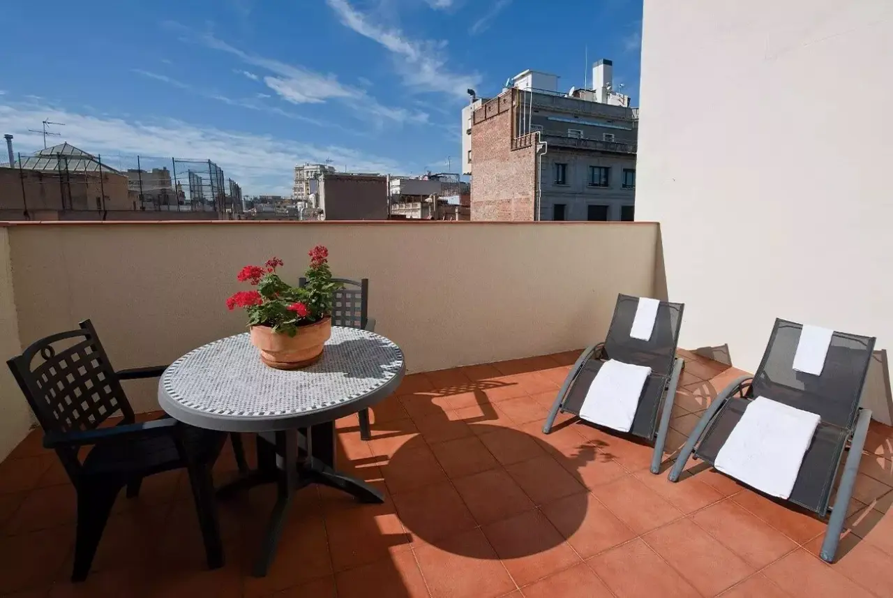 Terraza exterior con suelo de terracota, mesa redonda con silla, maceta con flores rojas y dos tumbonas con toallas