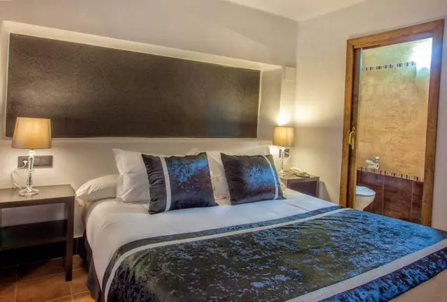 Vista de una junior suite con cama grande vestida en blanco y azul, cabecero oscuro iluminado, mesitas de noche