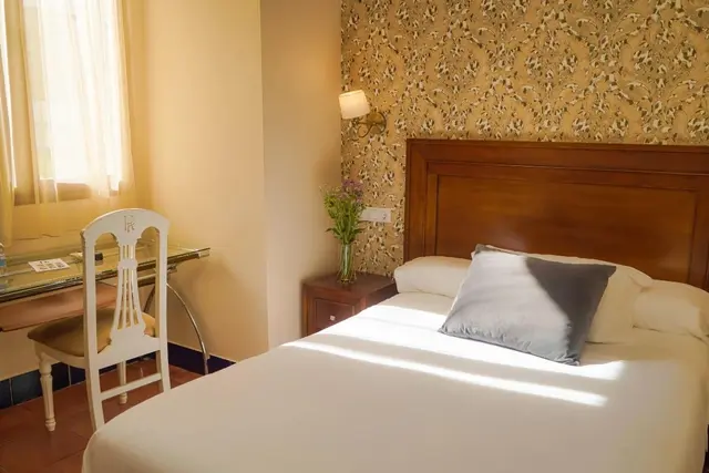 Habitación con cama doble, cabecero de madera, mesita de noche con jarrón de flores, escritorio con silla blanca