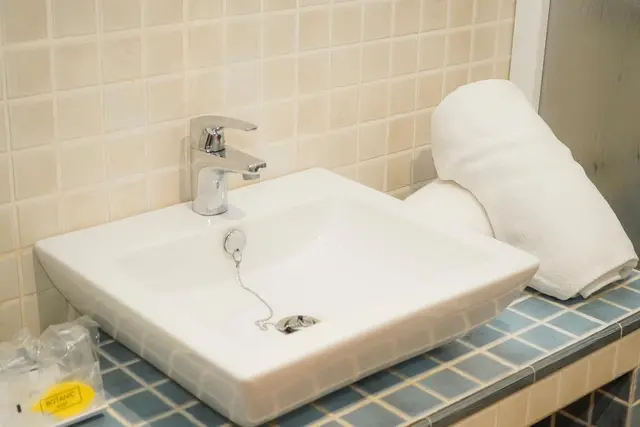 Lavabo cuadrado blanco sobre encimera de azulejos azules, con grifo cromado, desagüe con cadena y toallas blancas