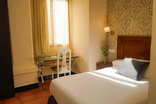 Vista de una junior suite con cama doble, escritorio con silla blanca, ventana con cortinas y papel pintado floral