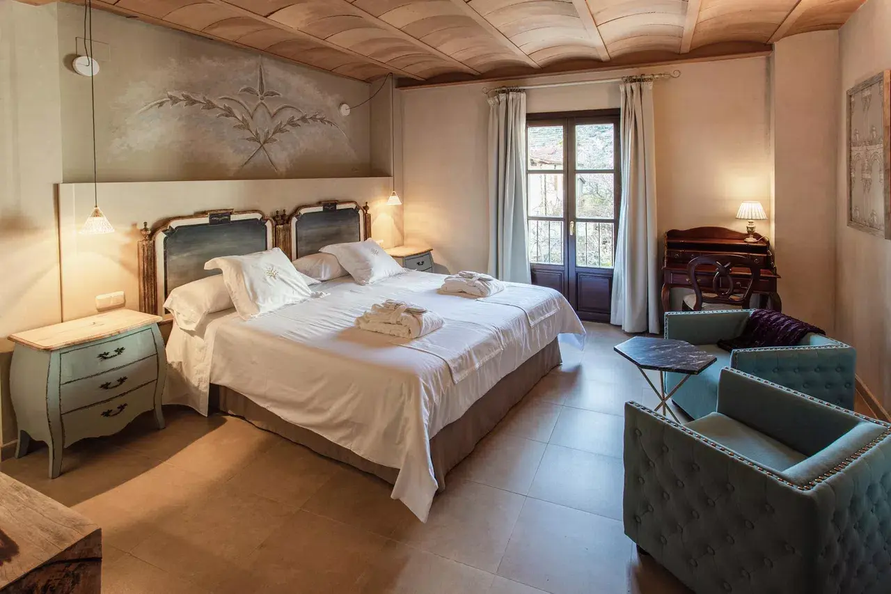 Habitación con cama doble grande vestida de blanco, dos mesitas de noche, cabecero con mural decorativo, techo