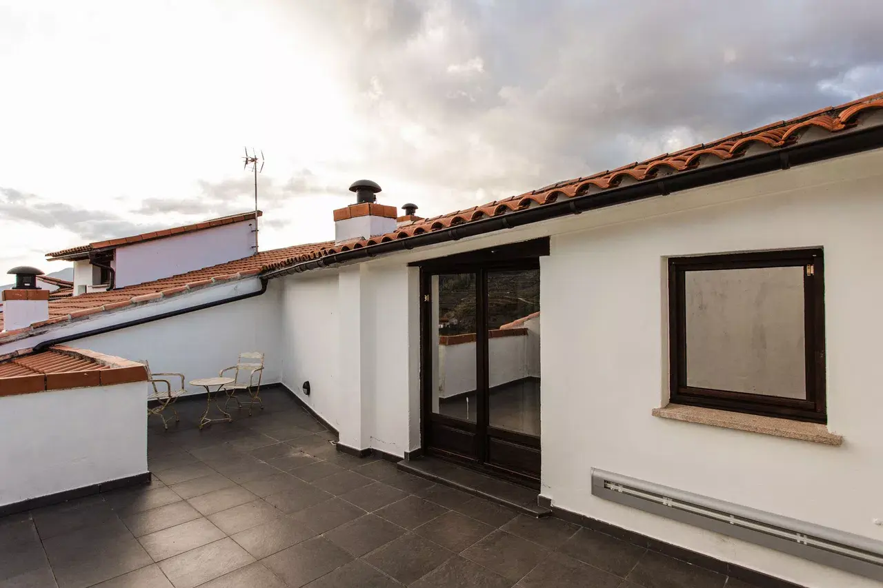 Terraza exterior de un edificio con suelo de baldosas oscuras, paredes blancas, tejado de teja árabe, mesa y sillas