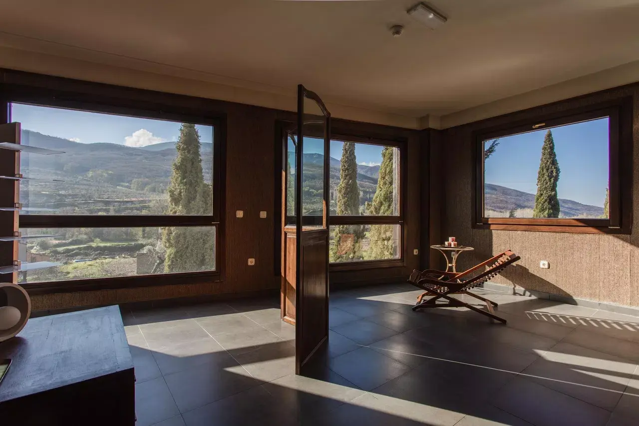 Interior de una sala amplia con suelo de baldosas oscuras, grandes ventanales que muestran un paisaje de montañas