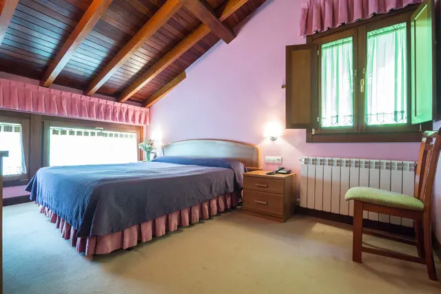 Habitación con cama doble, techo abuhardillado de madera, ventana con cortinas y silla junto a un radiador.