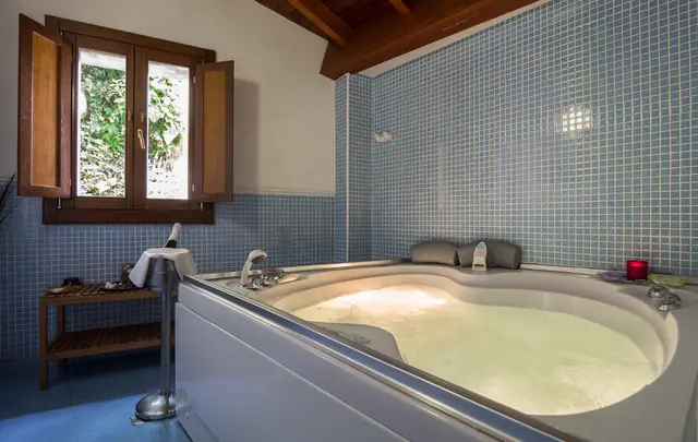 Bañera de hidromasaje iluminada en una habitación con paredes de azulejos azules y ventana de madera abierta.