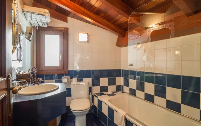 Baño con bañera, inodoro, lavabo y paredes de azulejos blancos y azules bajo techo de madera.