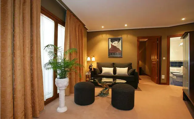 Salón de una habitación de hotel con sofá, mesa de centro, pufs, planta en maceta y puerta abierta a un baño con bañera.