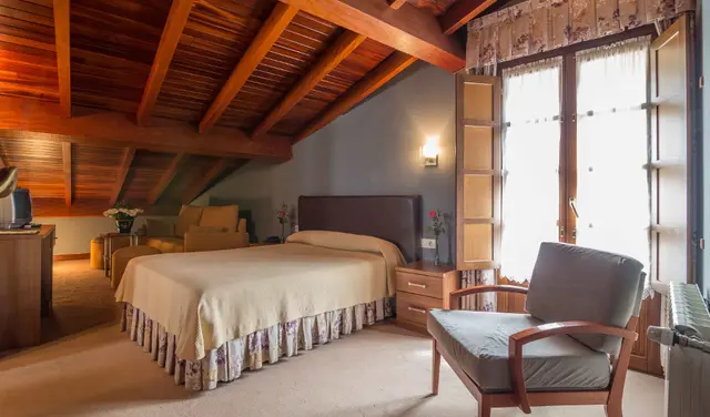 Habitación doble con cama grande, techo abuhardillado de madera, butaca y gran ventana con cortinas.