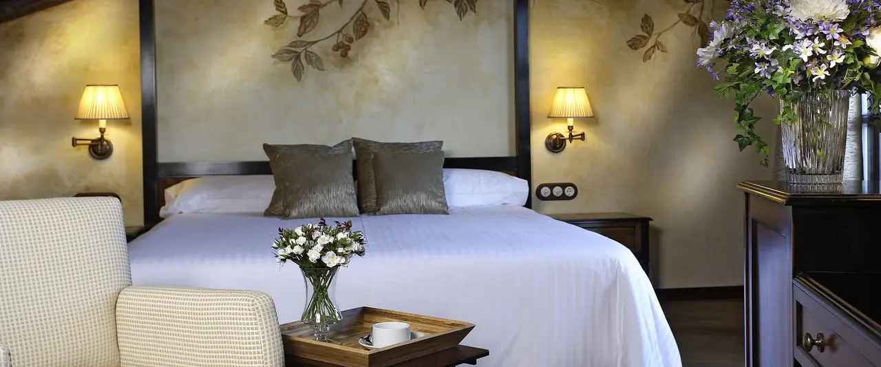 Habitación con cama doble grande vestida de blanco, cabecero oscuro, dos almohadas decorativas grises, mesitas
