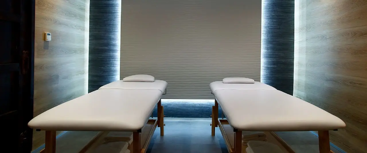 Dos camillas de masaje paralelas con colchones blancos y almohadas, colocadas en una sala con paredes de piedra
