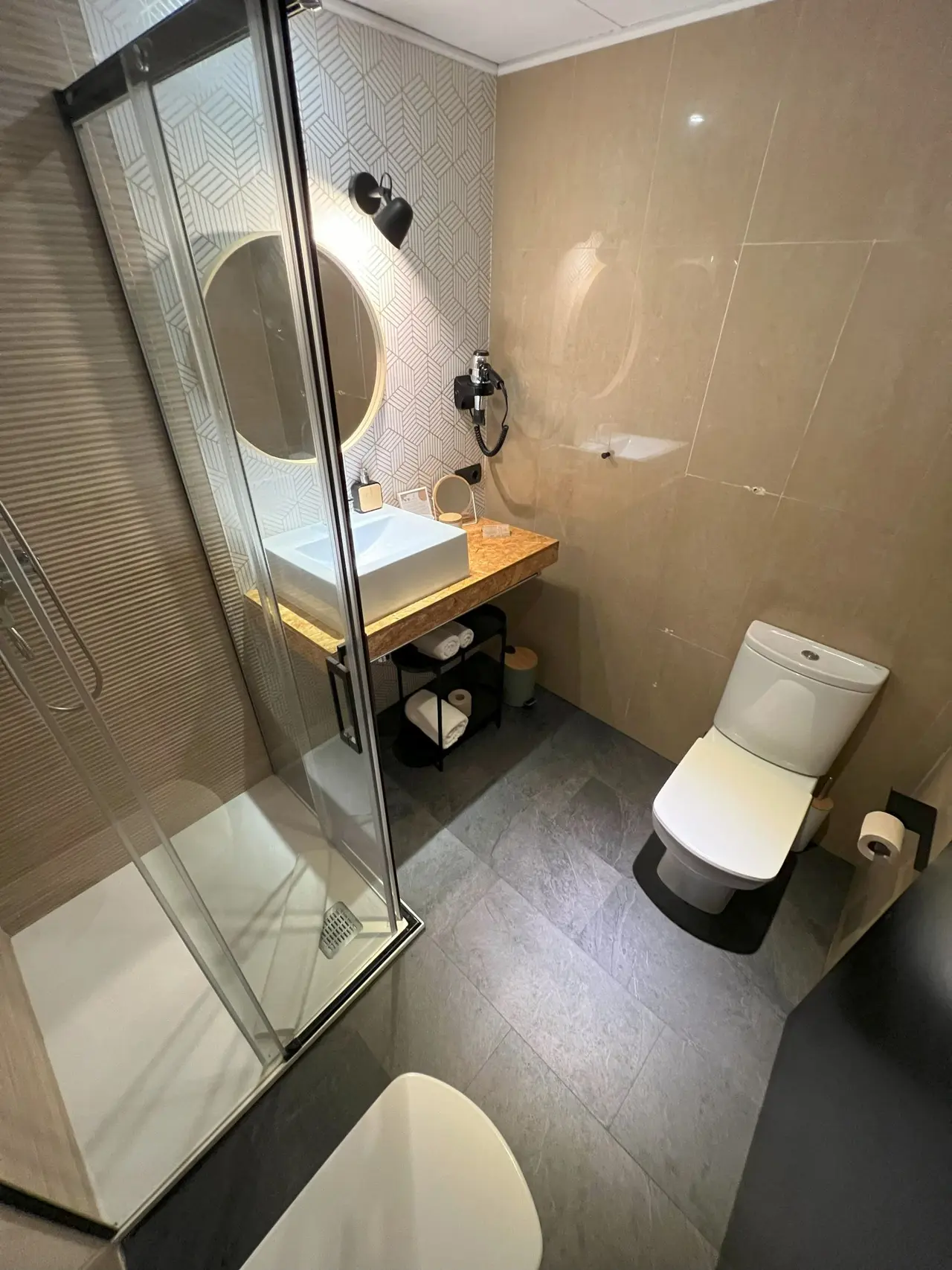 Baño de hotel con ducha acristalada a la izquierda, lavabo rectangular blanco sobre encimera de madera con espejo