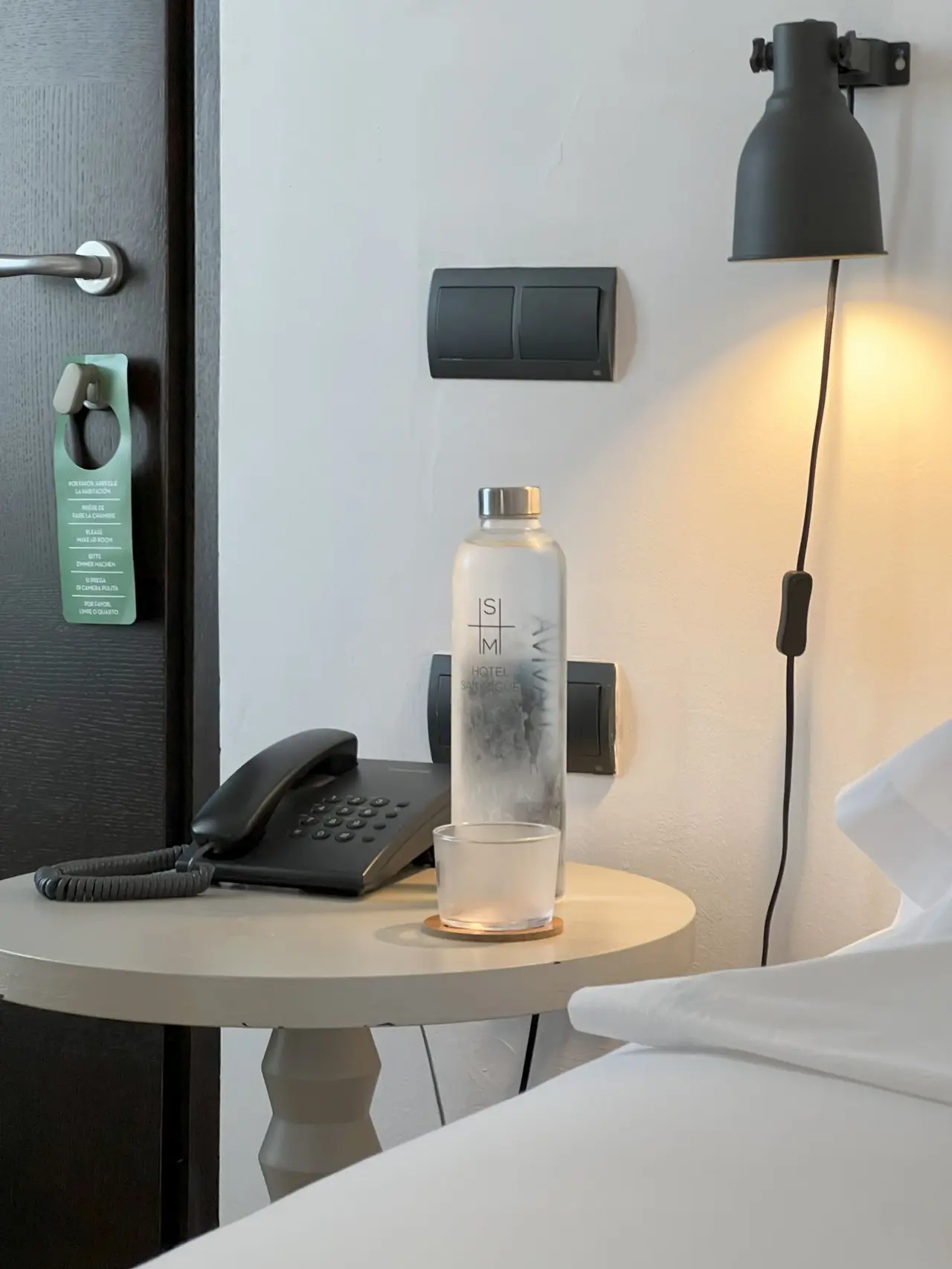 Mesa redonda junto a la cama en una habitación de hotel, con un teléfono fijo negro, una botella de agua de vidrio