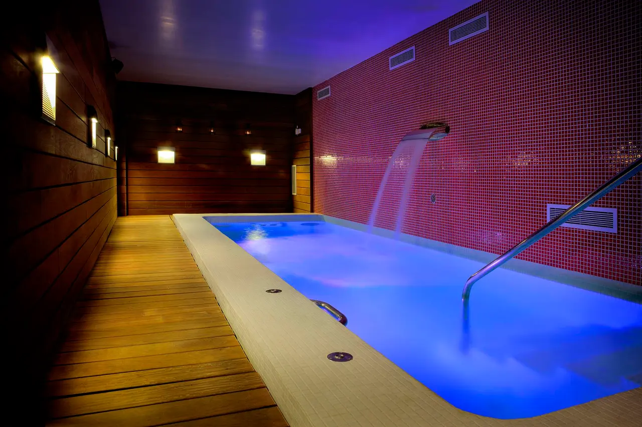 Interior de un spa privado con una piscina rectangular iluminada en azul, rodeada de suelo de madera, paredes