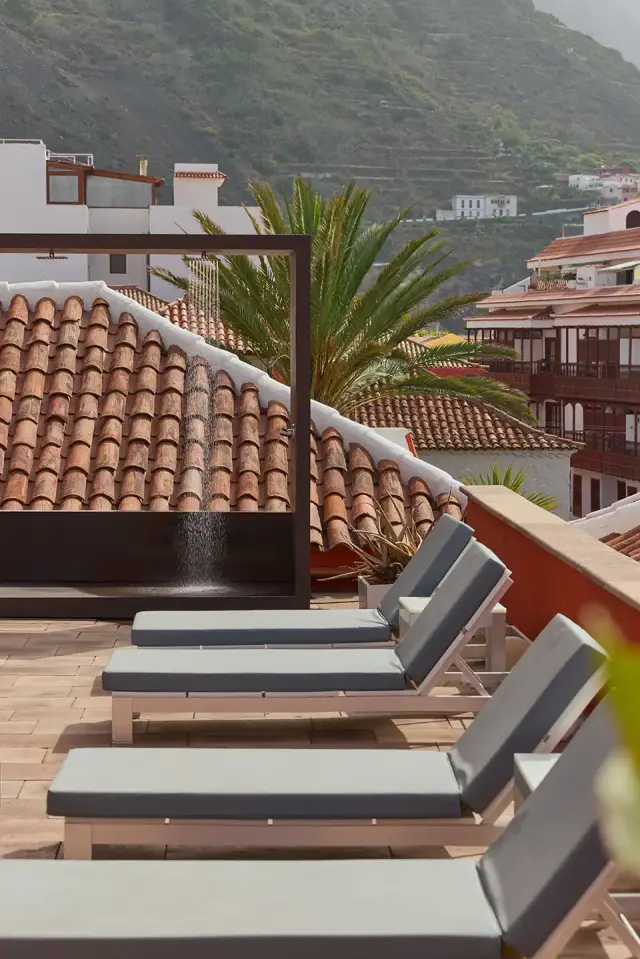 Terraza exterior con varias tumbonas en primer plano, una ducha al aire libre con agua cayendo y vistas a los tejados
