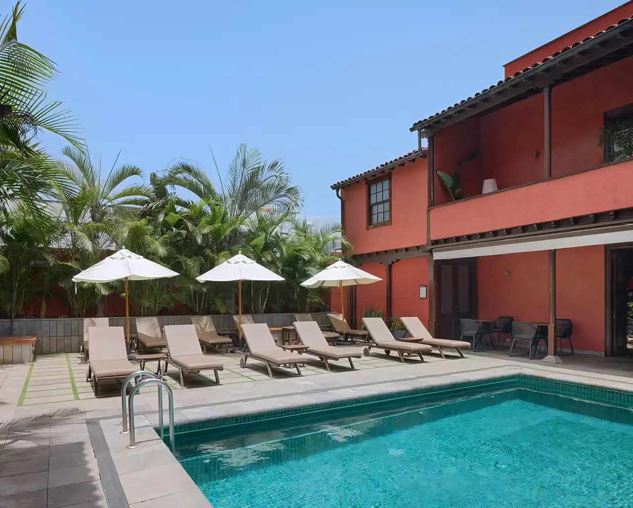 Vista de una piscina rectangular con agua turquesa en un patio exterior. Alrededor hay varias tumbonas con sombrillas