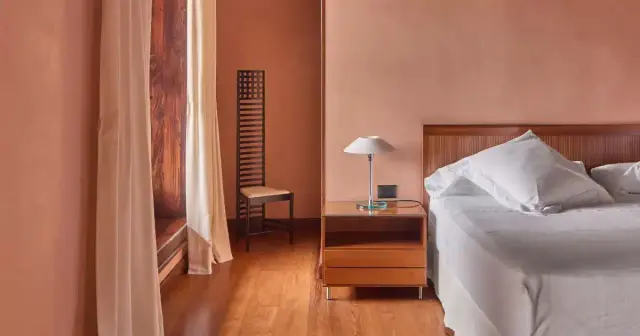 Interior de una suite con cama doble vestida de blanco, mesita de noche de madera con lámpara, silla alta de diseño