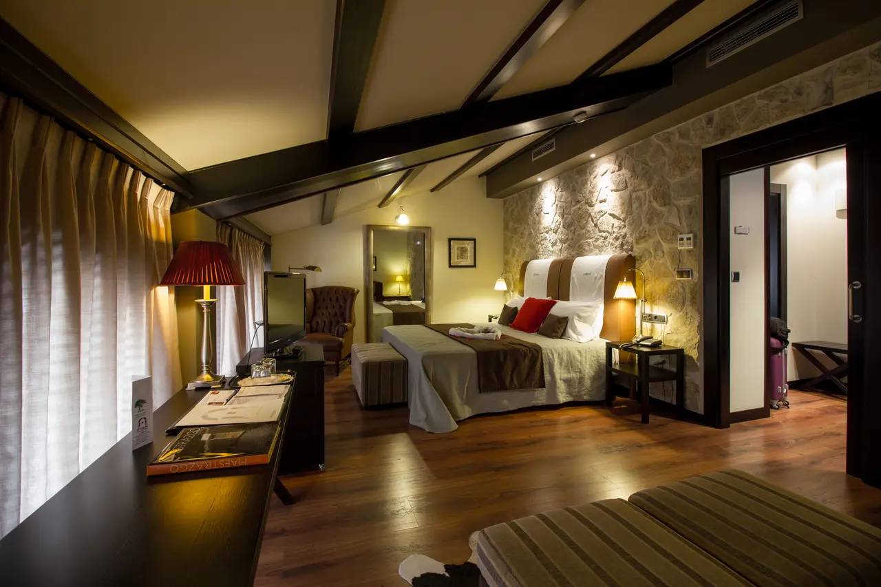 Vista de una habitación de hotel con cama doble, cabecero de piedra, vigas de madera en el techo, escritorio