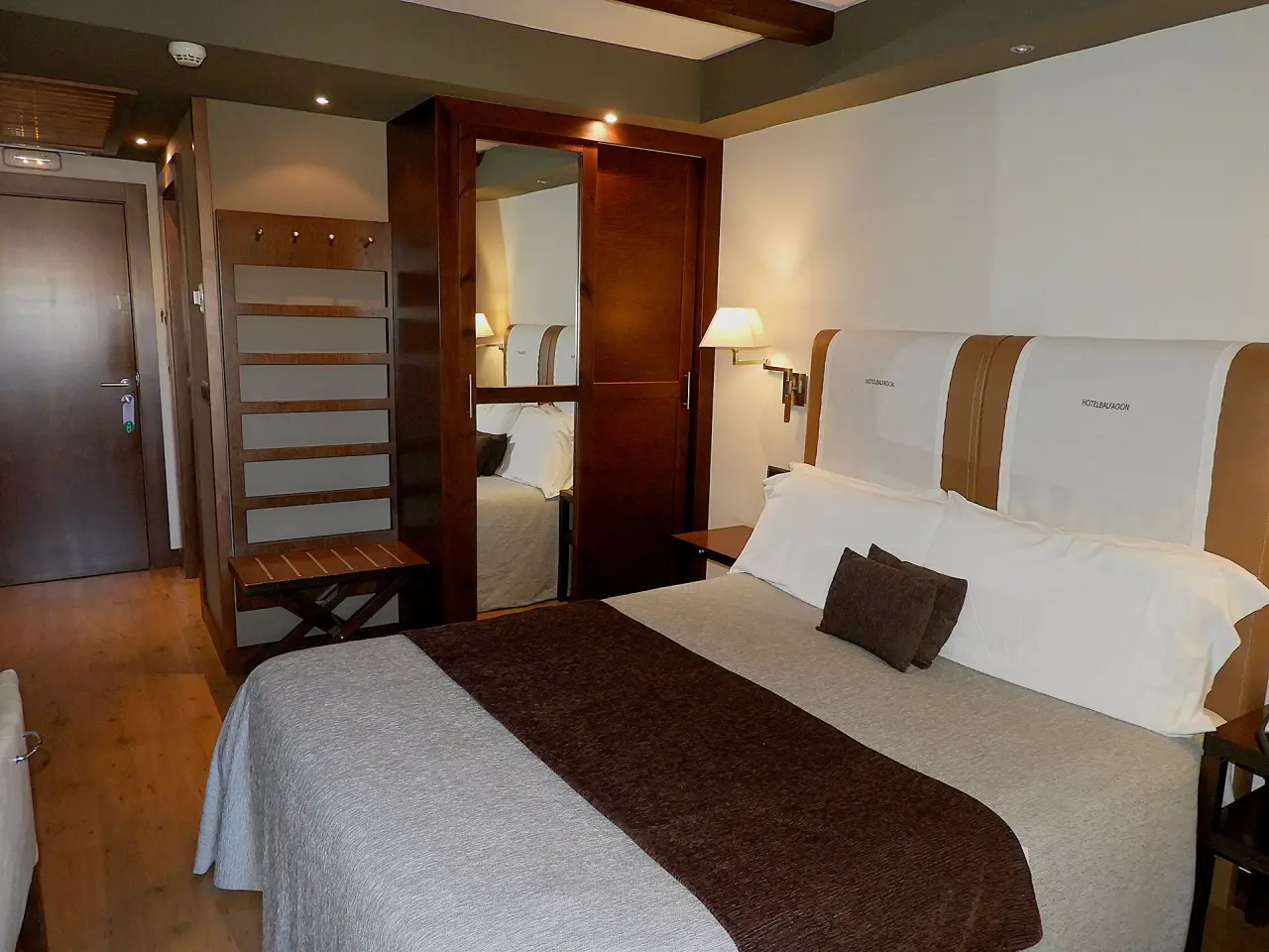 Habitación de hotel con cama doble grande vestida con colcha clara y runner marrón, cabecero alto blanco y marrón