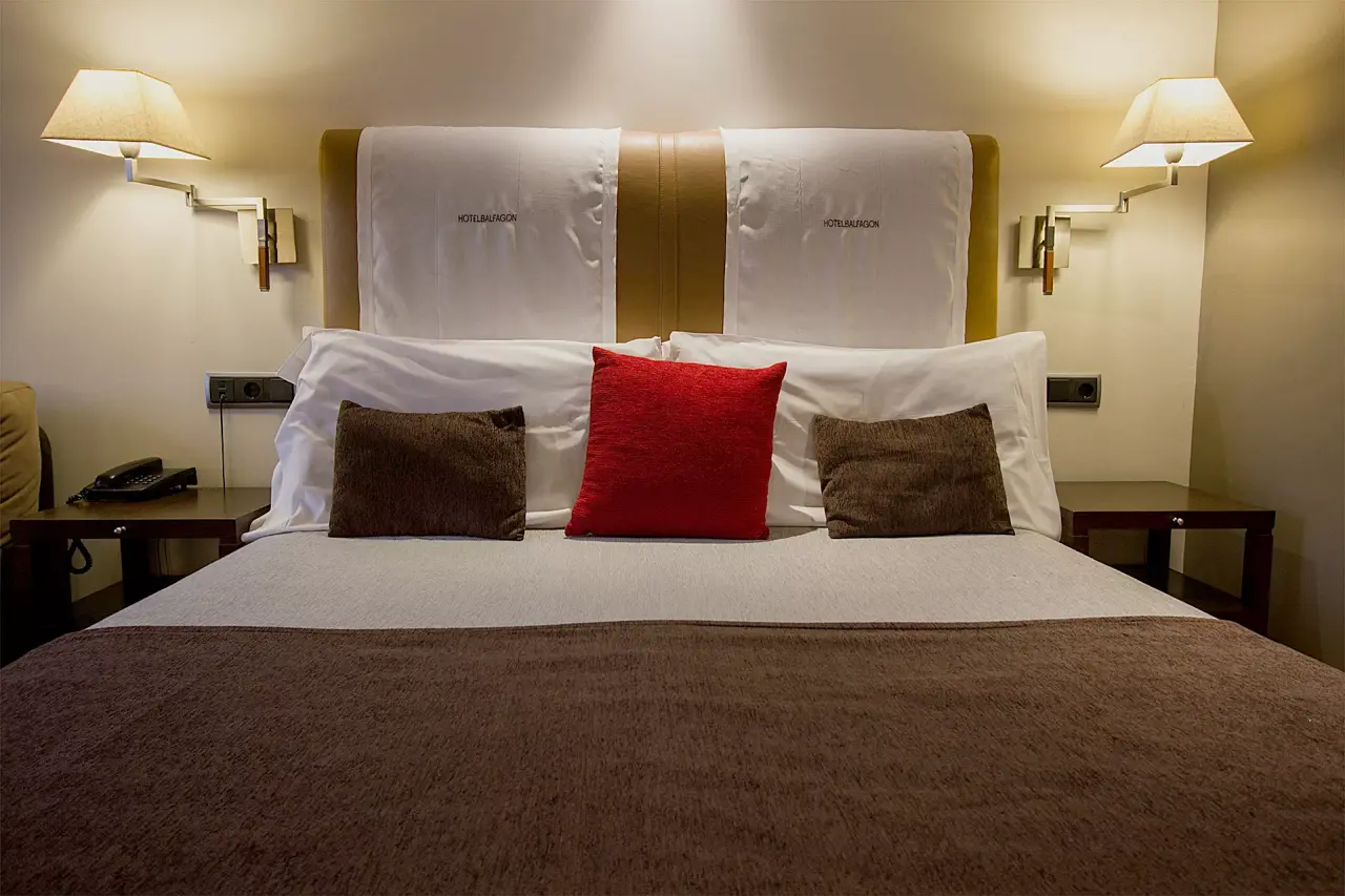 Vista frontal de una cama matrimonial con cabecero acolchado en tonos beige y blanco, dos almohadas grandes blancas