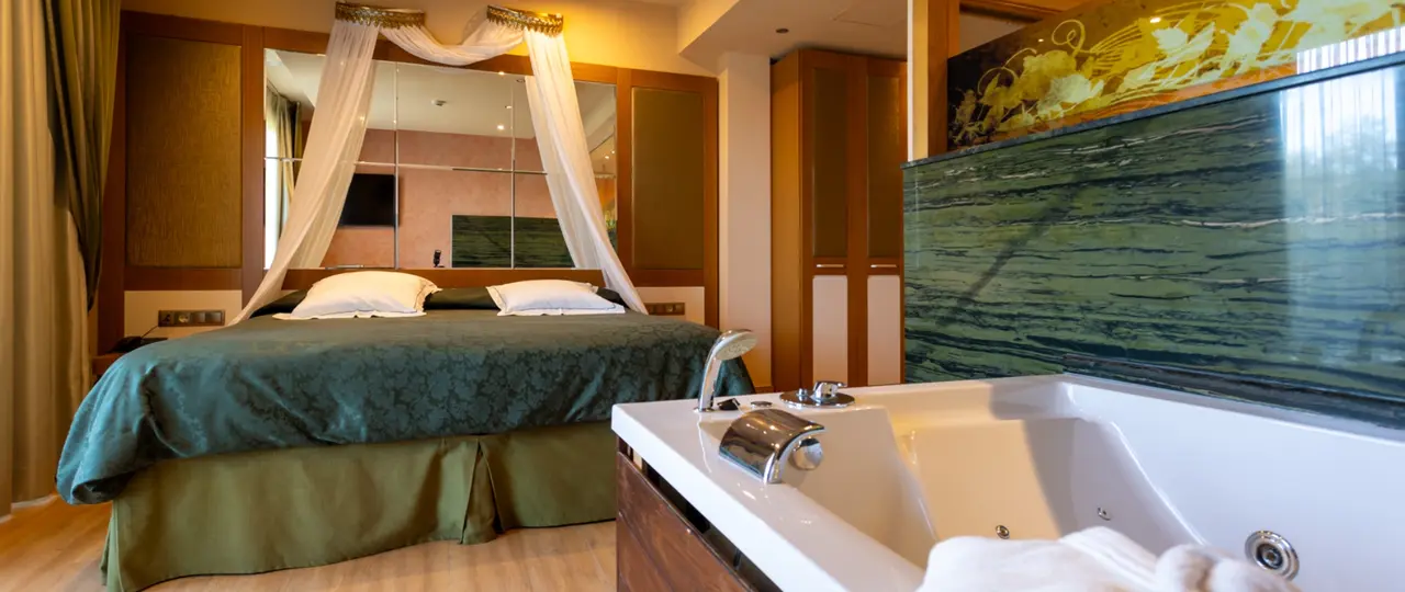 Habitación de hotel con cama de matrimonio con dosel y cortinas blancas, y bañera de hidromasaje blanca en primer