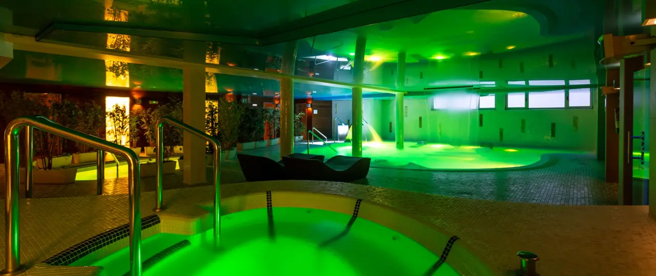 Vista de un spa interior con una bañera de hidromasaje en primer plano iluminada en verde, rodeada de un área