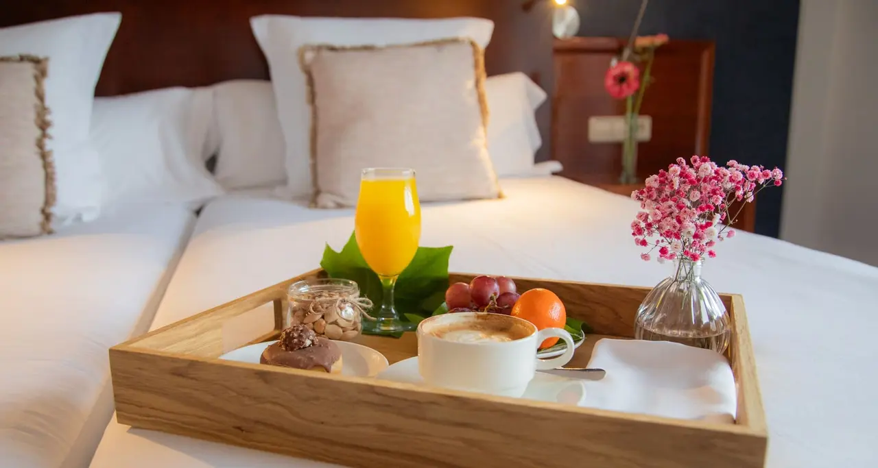 Bandeja de desayuno con café, zumo de naranja, frutas y flores sobre una cama blanca en una habitación de hotel.