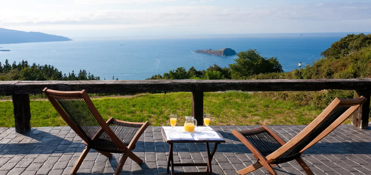 Vista panorámica al mar desde una terraza con dos tumbonas de madera y una mesa con vasos de zumo de naranja, rodeada