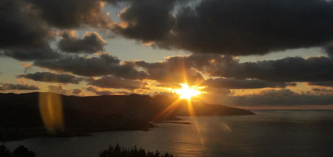 Vista panorámica de un atardecer sobre el mar y montañas, con el sol brillando entre nubes oscuras y un reflejo