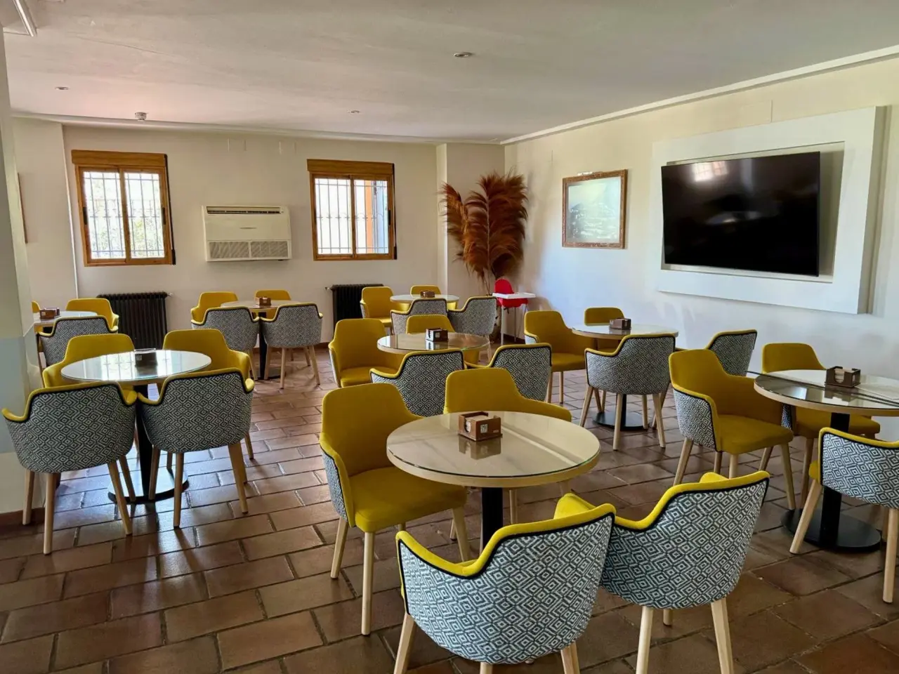 Espacioso salón con mesas redondas, sillas amarillas y tapizadas, suelo de terracota y televisión en la pared.