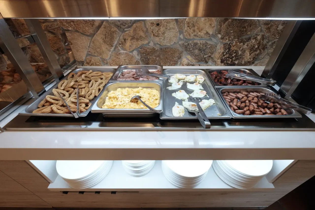 Buffet de desayuno con bandejas de churros, huevos revueltos, bacon, salchichas y platos apilados debajo.