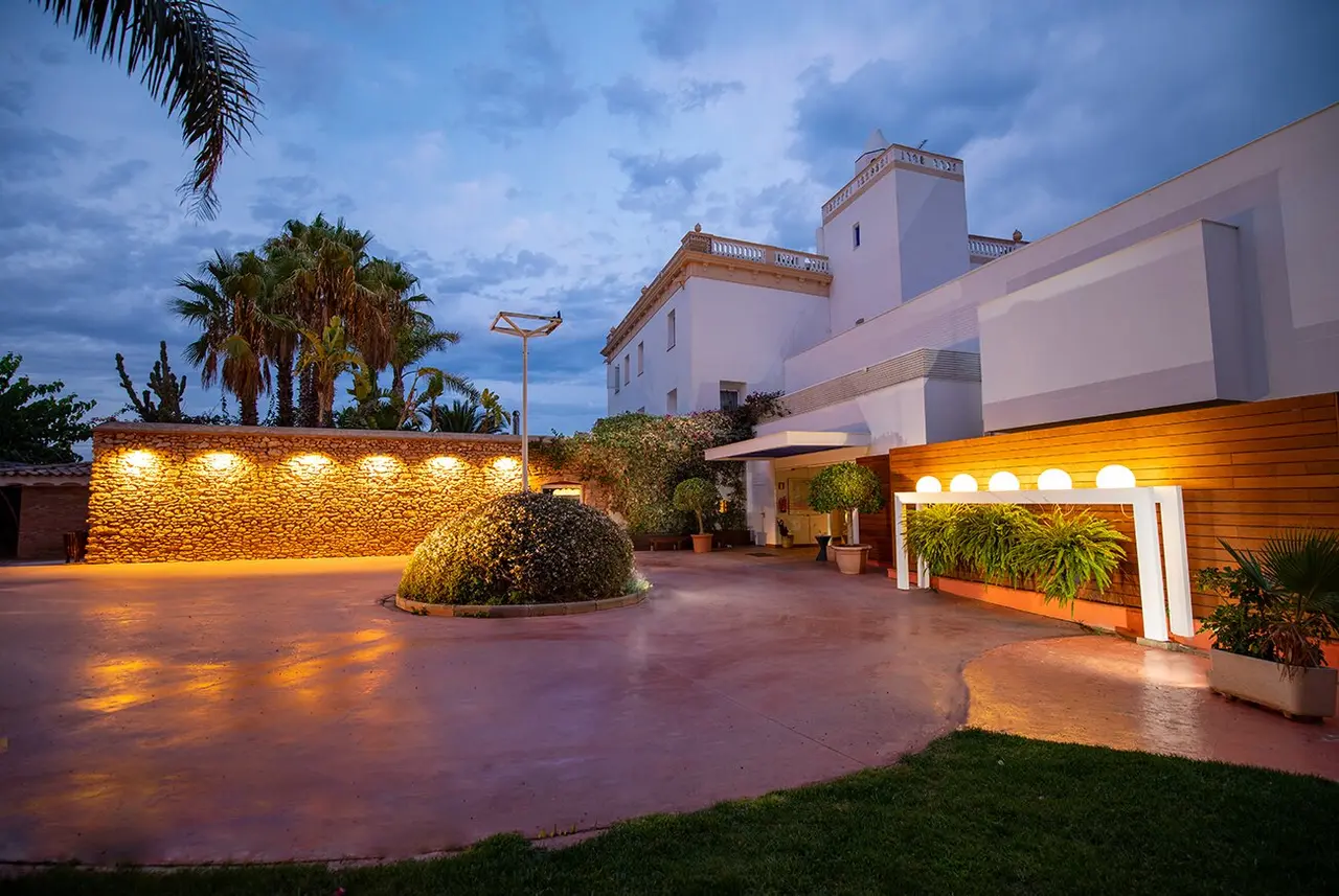 Vista exterior de un hotel al atardecer con fachada blanca, jardín con palmeras, setos iluminados, pavimento