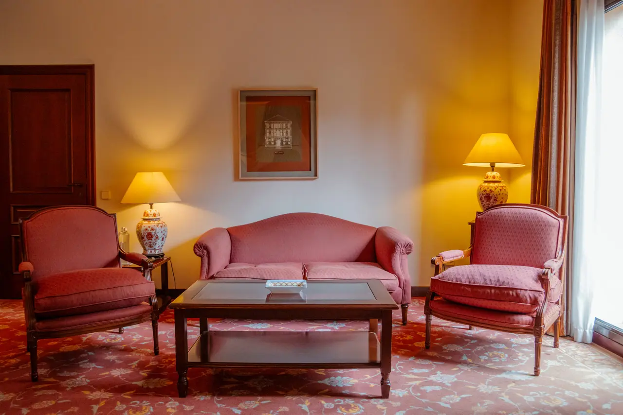 Salón de una suite con sofá de tres plazas y dos sillones tapizados en rosa, mesa de centro de madera, dos lámparas