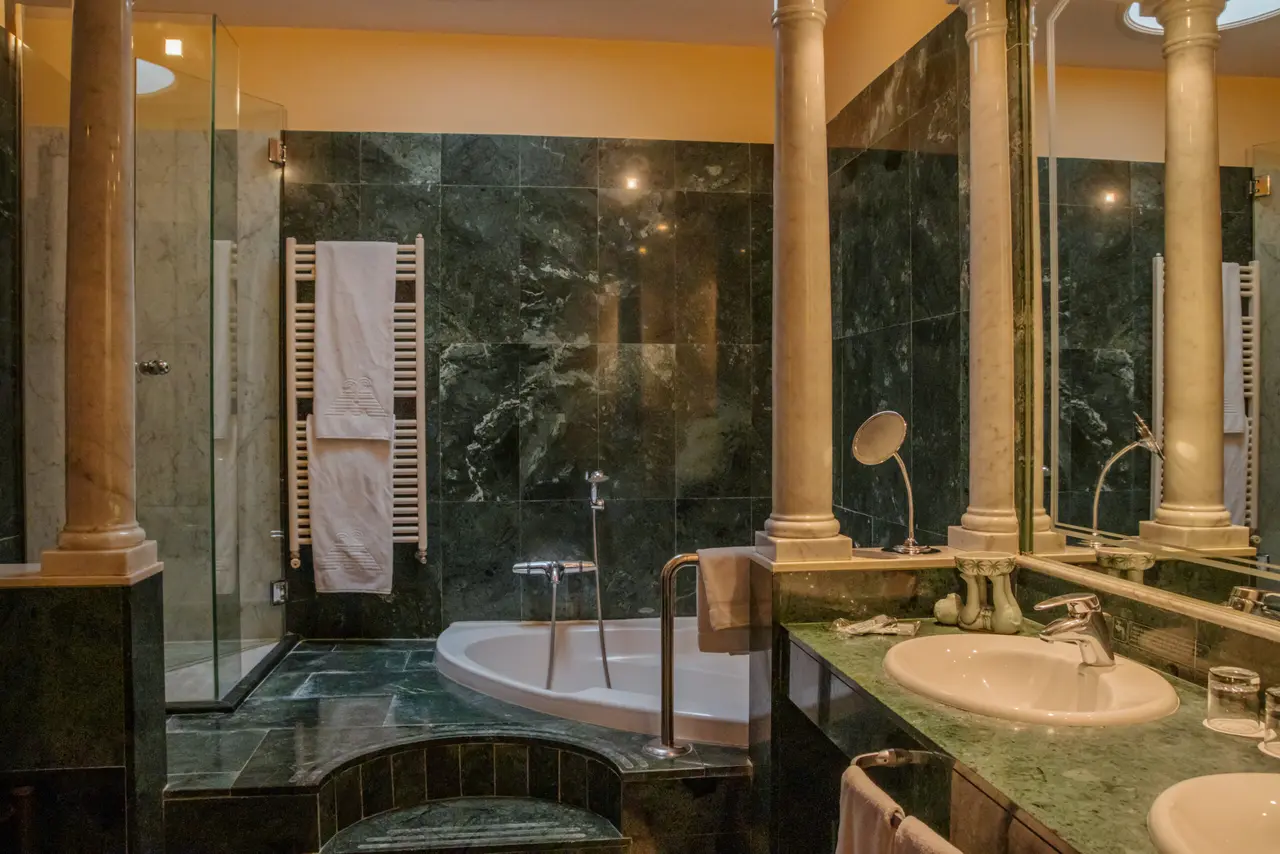 Baño de suite con bañera de hidromasaje blanca empotrada en el suelo, rodeada de paredes de mármol verde oscuro, ducha