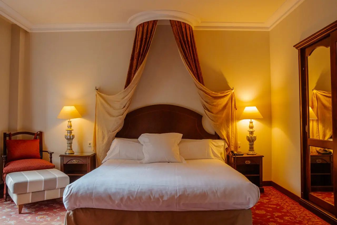 Vista de una suite con cama king size con dosel de cortinas, dos mesitas de noche con lámparas encendidas, sillón
