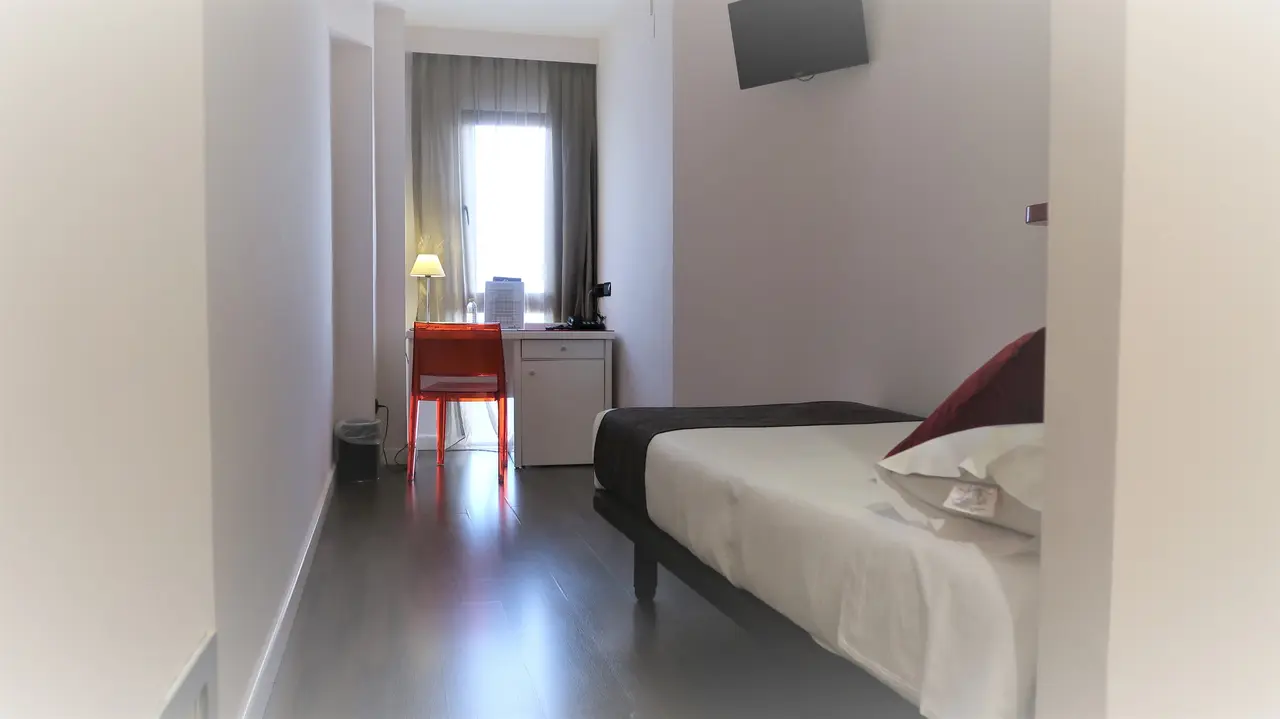 Vista de una junior suite con cama doble, escritorio con silla roja, ventana con cortinas y televisor en la pared.