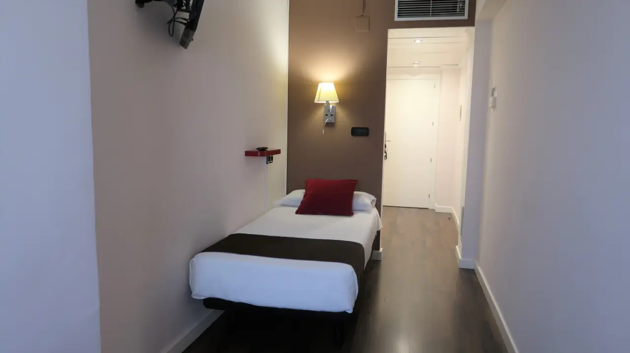 Vista de una habitación de hotel con una cama individual vestida con sábanas blancas, manta marrón y cojines rojos.