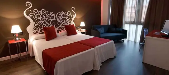 Vista de una junior suite con cama doble grande, cabecero blanco decorativo, almohadas rojas, mesitas de noche