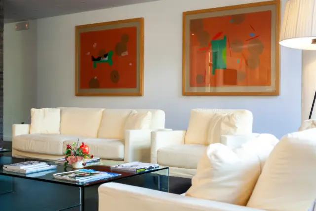 Salón de suite con sofás blancos, mesa de centro con libros y flores, y dos cuadros abstractos en la pared.