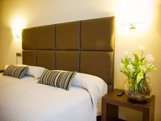 Vista de una cama doble con cabecero acolchado marrón, almohadas y cojines decorativos, mesita de noche con jarrón