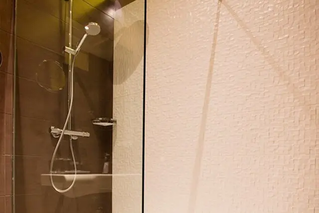 Vista de una ducha con mampara de vidrio, grifo y alcachofa de ducha metálicos, y pared lateral con azulejos blancos