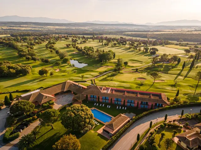 Vista aérea del Hotel TorreMirona Golf & Spa rodeado de un extenso campo de golf con lagos, árboles y montañas al fondo.